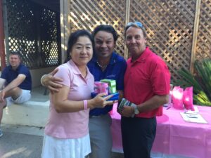 A smiling trio poses outdoors, holding gift bags, with a pink table covered in prizes behind them.