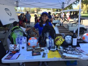 Young woman in a blue shirt and cap stands smiling behind a table filled with soccer balls, backpacks, and raffle items under a canopy at an outdoor event.