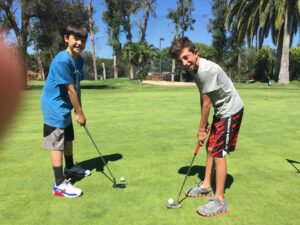 Two boys smiling while playing mini-golf on a sunny day, with putters poised over golf balls on a green course surrounded by trees.