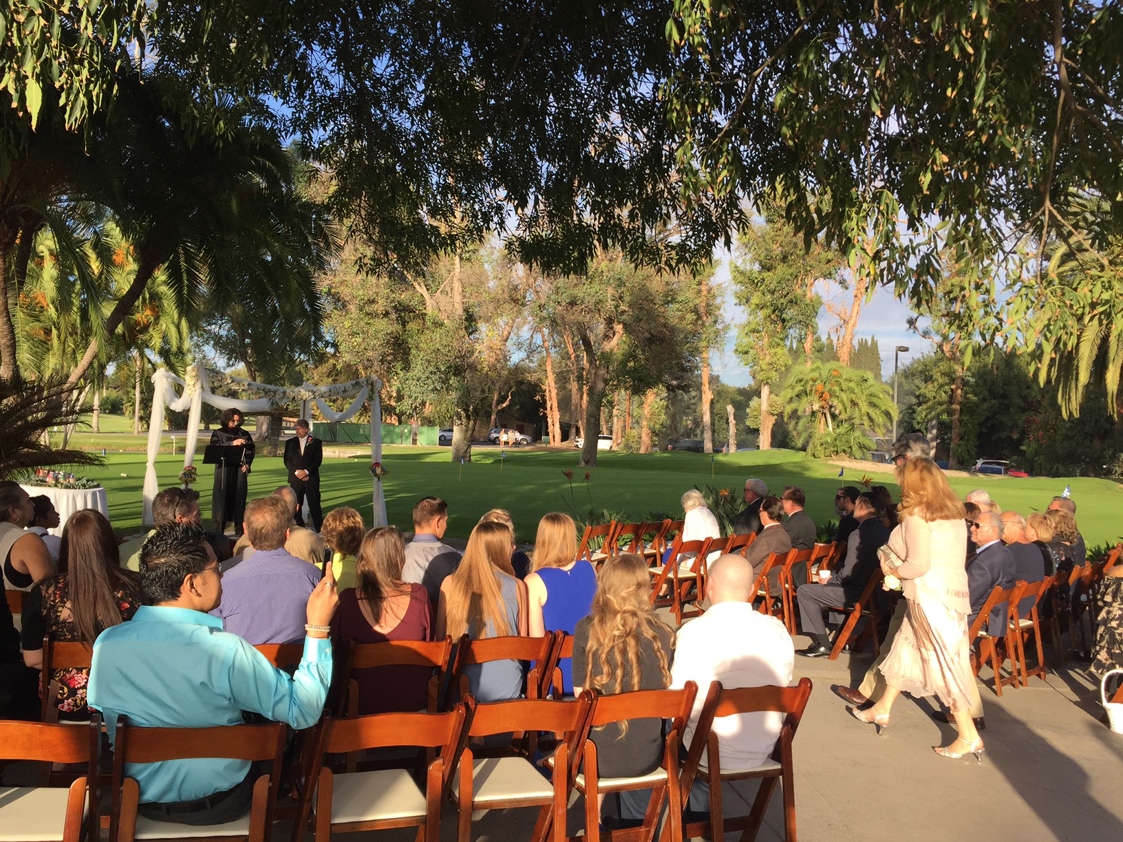 Outdoor wedding ceremony in a sunlit garden with guests seated on wooden chairs. A decorated arch in front and trees in the background.