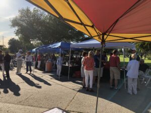 People gather under colorful tents at a sunny outdoor event. Groups converse and explore stalls, creating a lively, festive atmosphere.