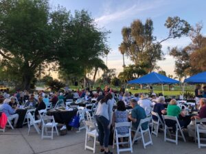 Outdoor social event at sunset with large group seated at round tables under blue canopies.
