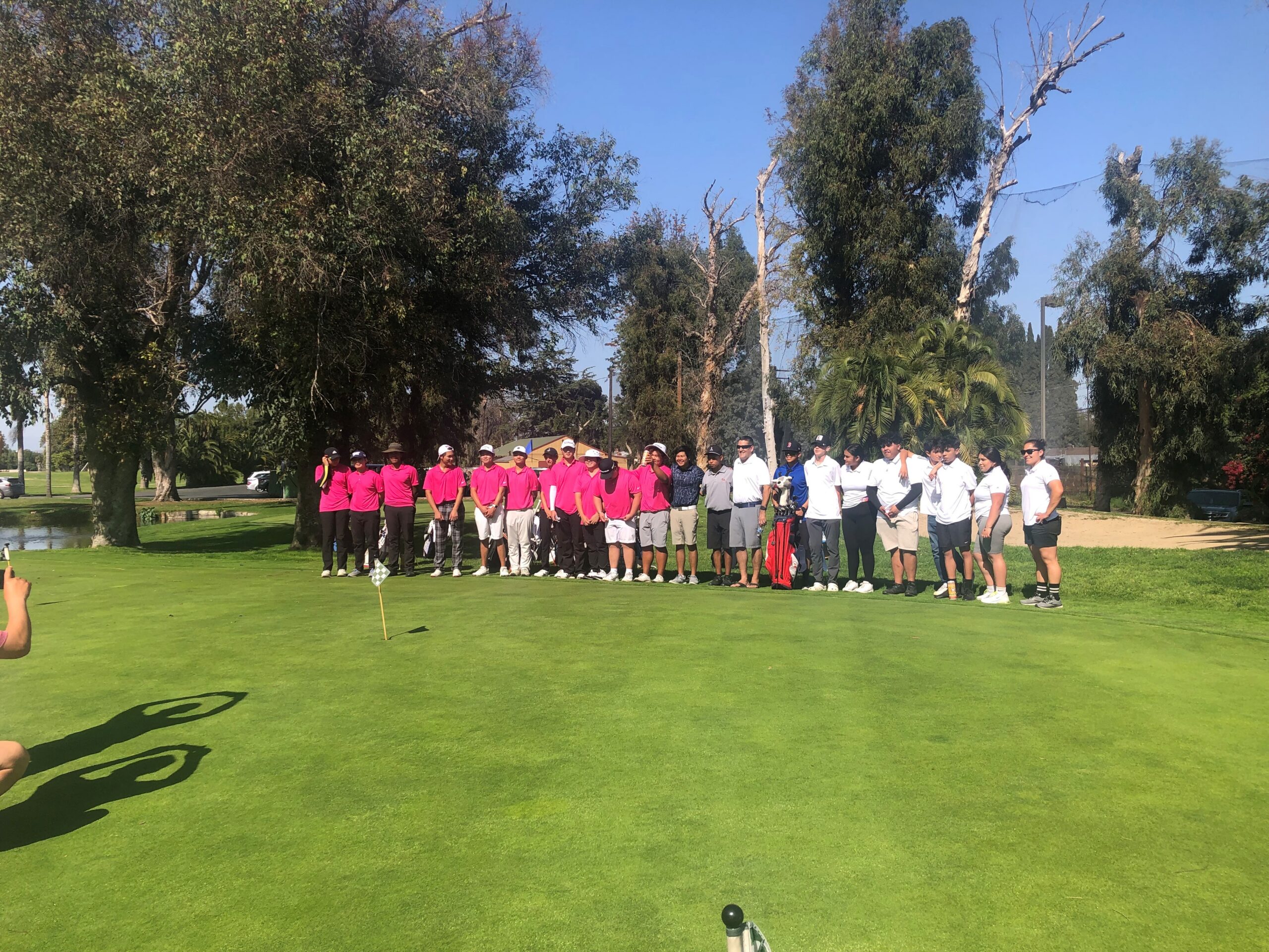 A group of people stands on a sunny golf course, divided into two teams wearing matching pink and white shirts. Trees and a small pond are in the background.