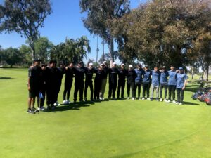 A group of people, dressed in golf attire, stand in a line on a golf course green, posing for a photo with trees and golf bags in the background.