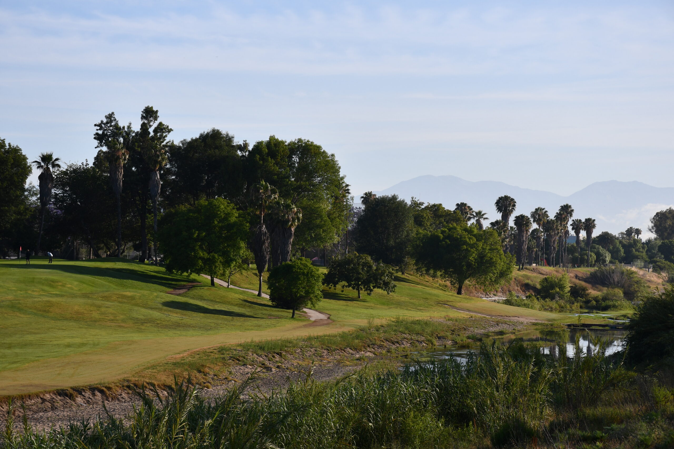 Scenic view of a golf course at sunrise, with lush green fairways, palm trees, a gentle river, and distant mountains under a clear blue sky.