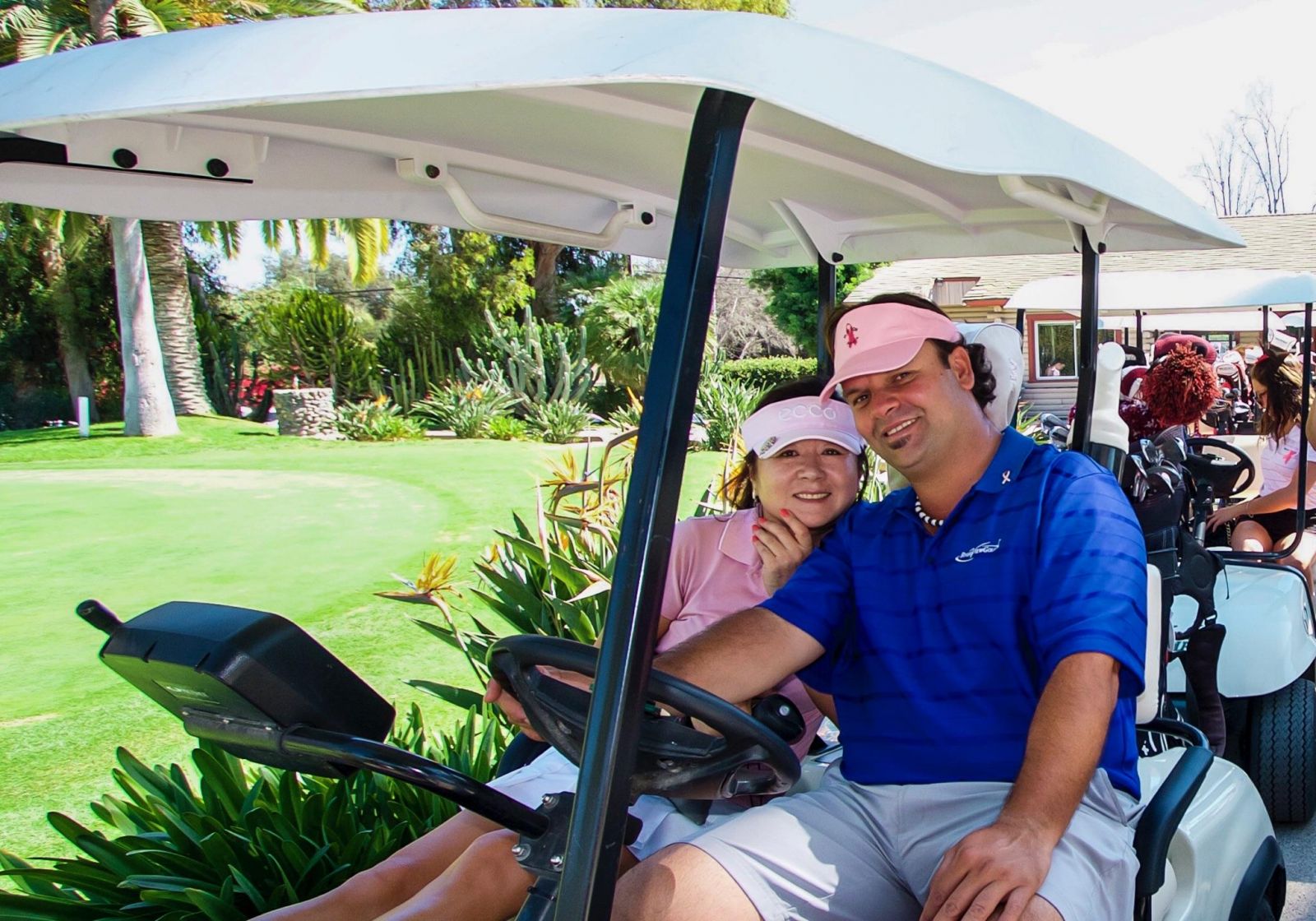 A man and woman smile while sitting in a golf cart. They're wearing casual golf attire, surrounded by lush greenery and palm trees.