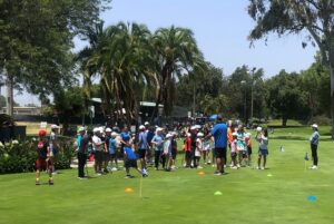 A group of children in colorful sports attire and white hats gather on a green field, listening to an instructor in blue under palm trees.