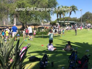 Children practicing golf on a sunny green course during a junior camp. Surrounded by trees and golf bags.