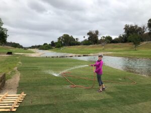 A person in a pink jacket waters grass with a hose near a tranquil river.