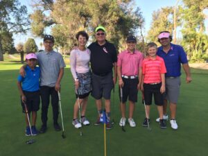 A group of six people pose on a golf course, smiling. They hold golf clubs and wear casual golf attire, including hats and pink accessories.