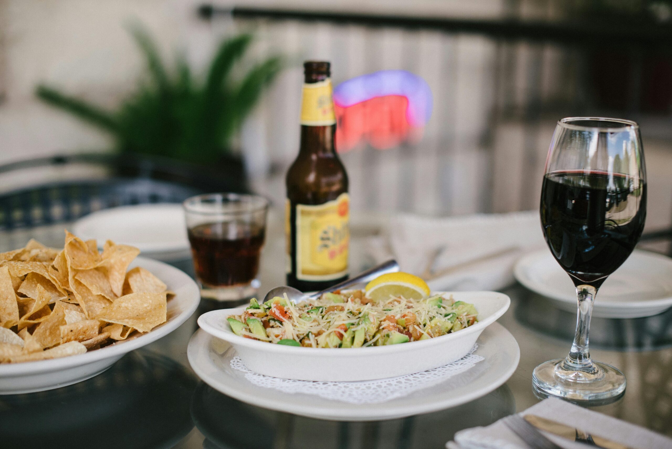 Glass table with a plate of salad topped with cheese next to tortilla chips. A bottle of beer, glass of red wine, and cola complete the setting.