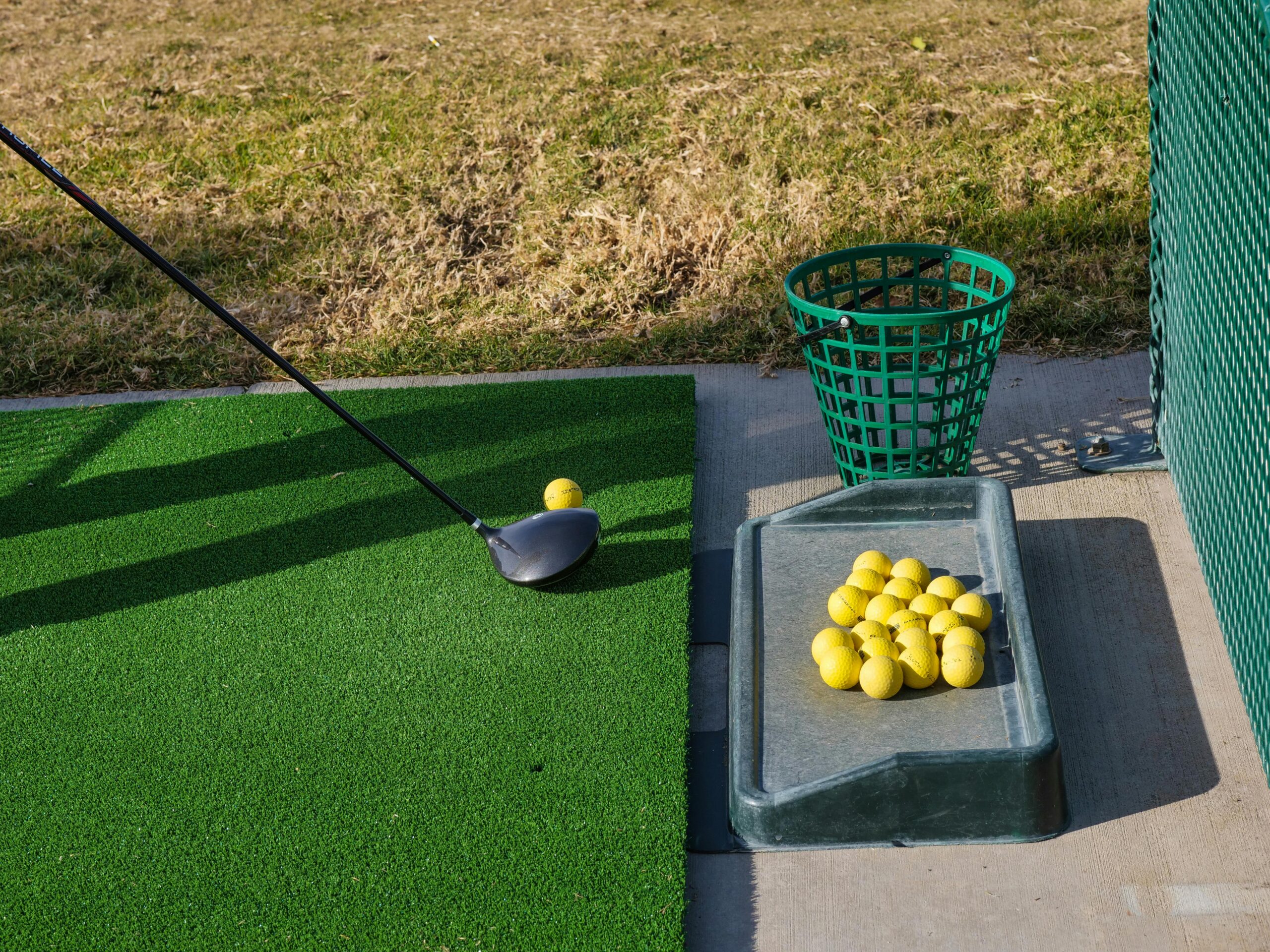 A golf club and yellow ball on green turf near a tray with more balls. A mesh basket is in the background, with grass lining the scene.