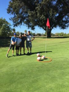 Four people stand on a sunny golf course green, smiling beside a flagstick. Soccer balls rest in a hoop near the hole, trees in the background.