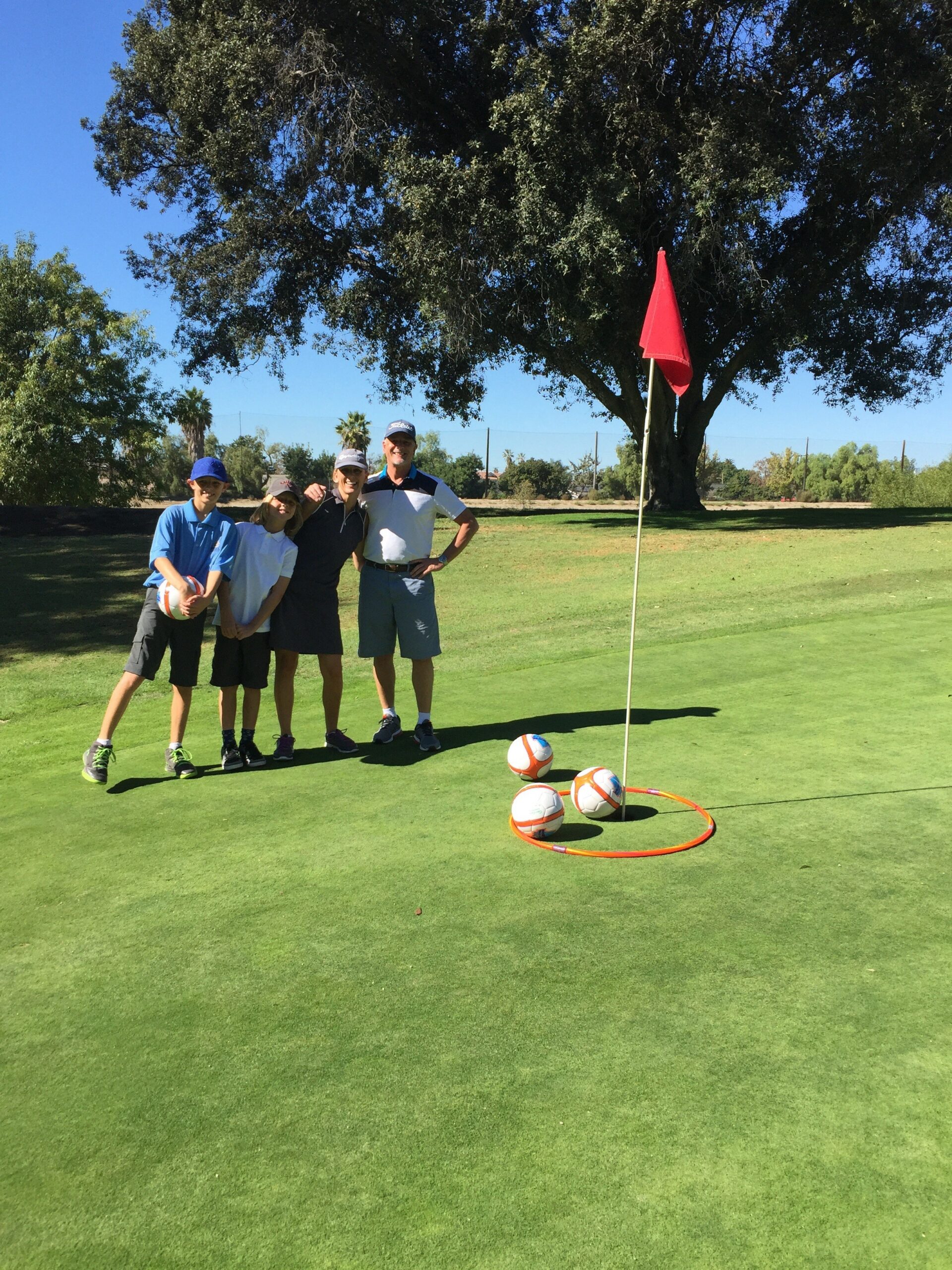 Four people stand on a sunny golf course green, smiling beside a flagstick. Soccer balls rest in a hoop near the hole, trees in the background.