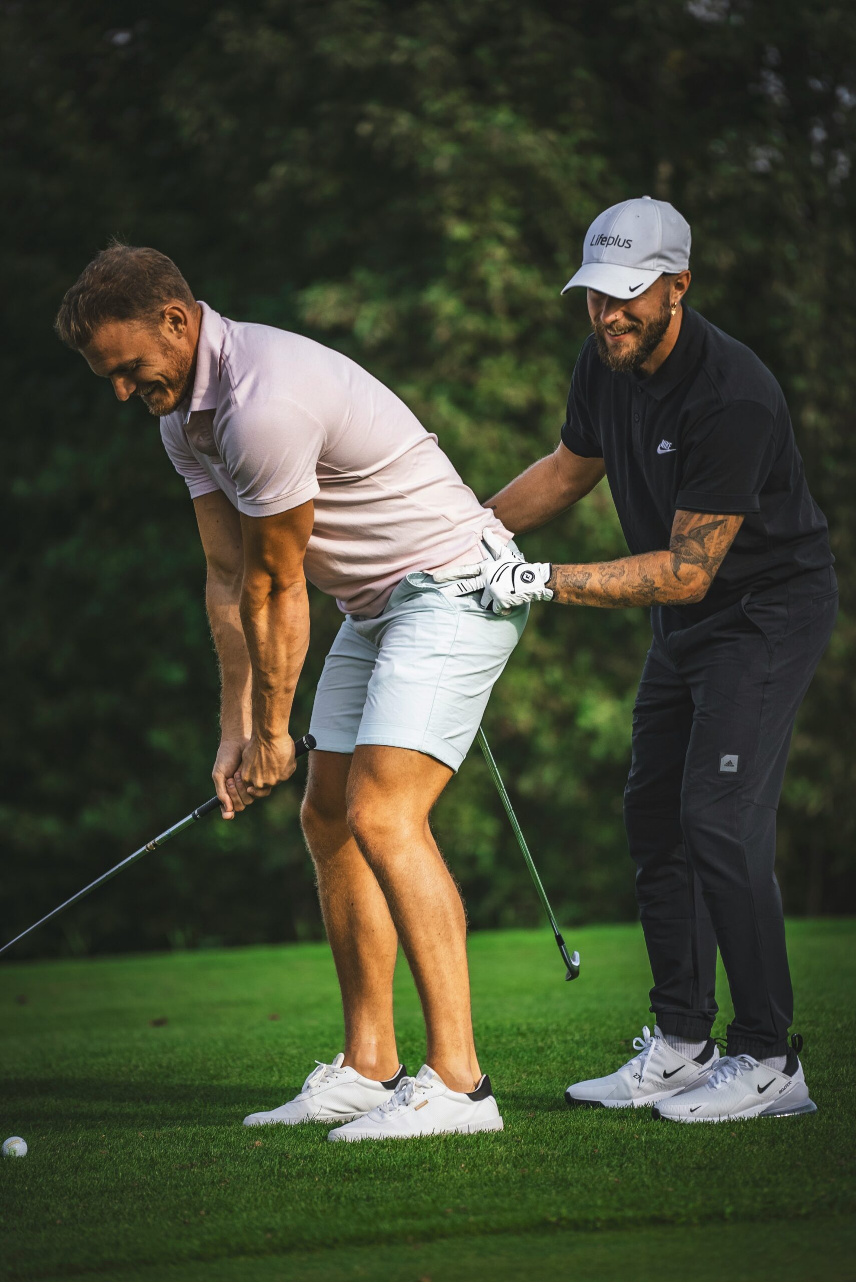 Two men on a golf course; one in a pink shirt and shorts is preparing to swing, while the other in black adjusts his posture.