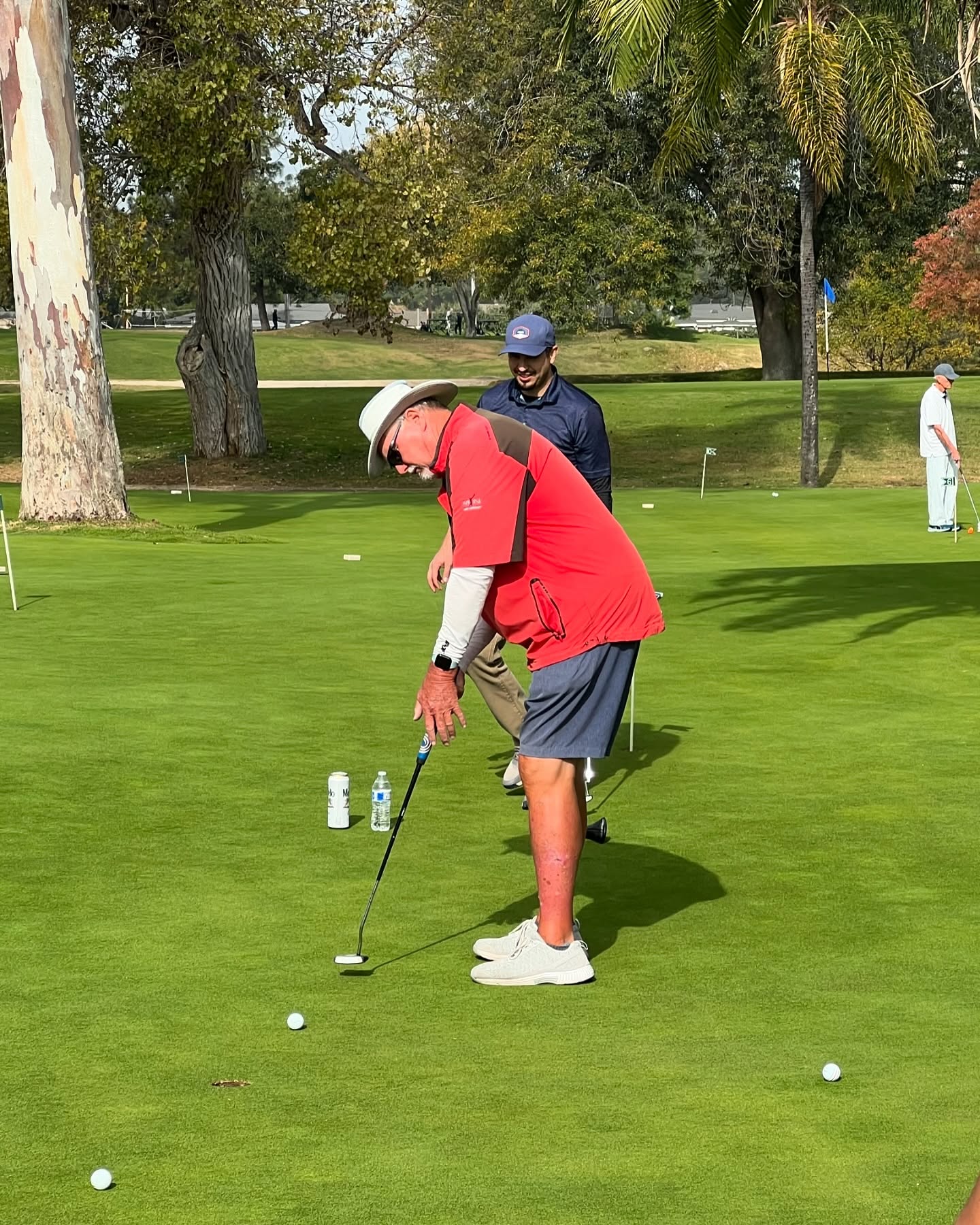 Golfer in a red shirt and white hat putts on a green, surrounded by trees. A fellow golfer watches in the background.