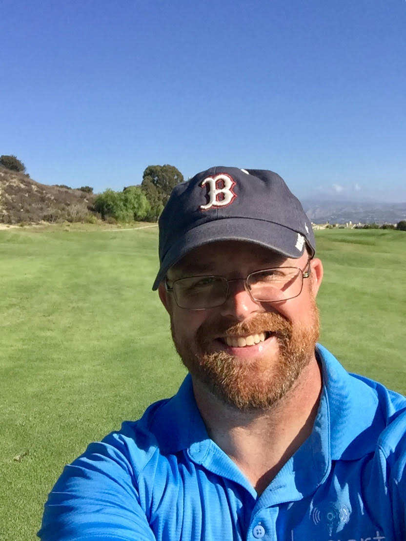 Man wearing a cap and glasses takes a selfie on a sunny golf course. He's smiling broadly, wearing a blue shirt with clear skies above.