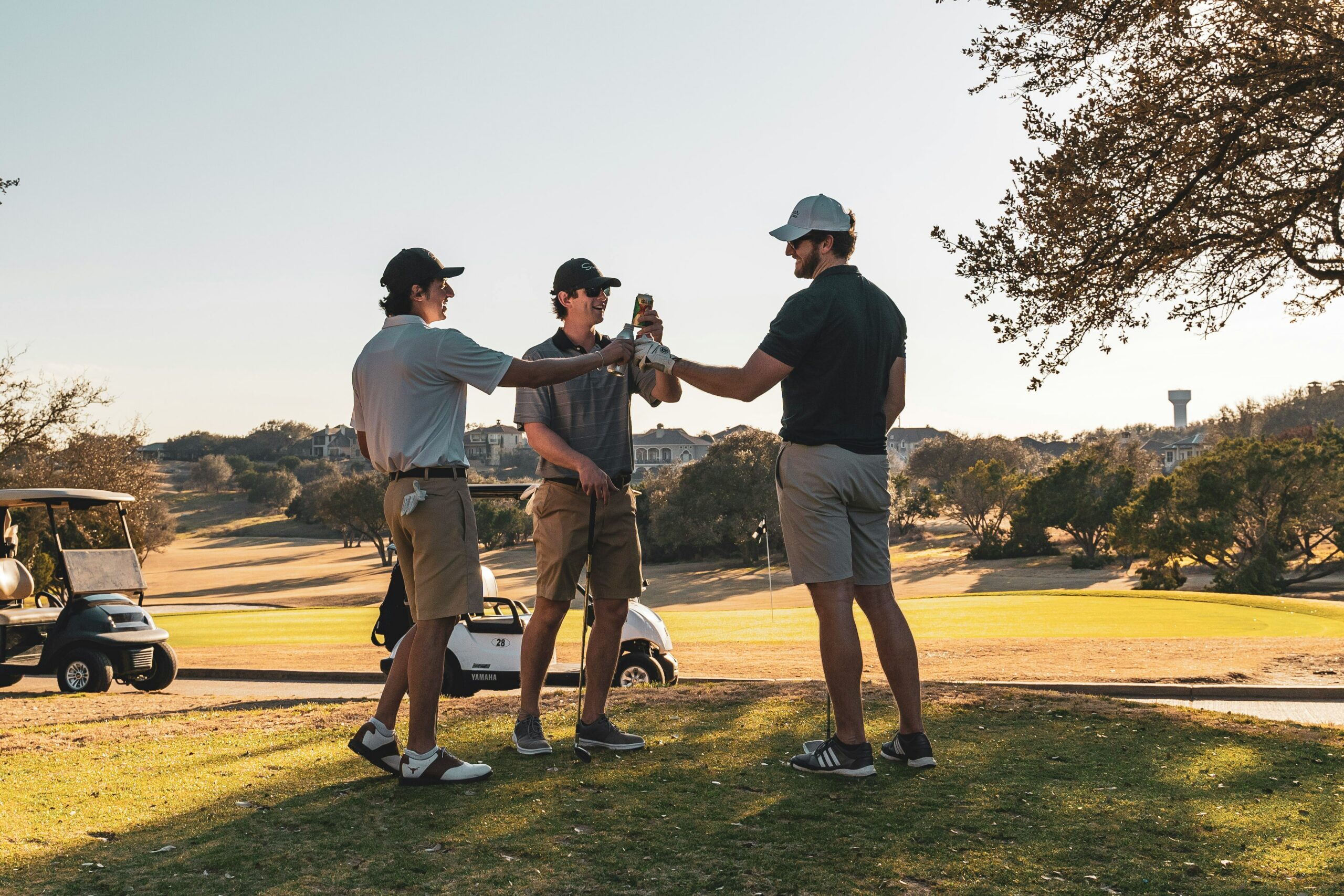 Three men in casual golf attire toast with bottled drinks on a golf course. A golf cart and trees are visible in the background,