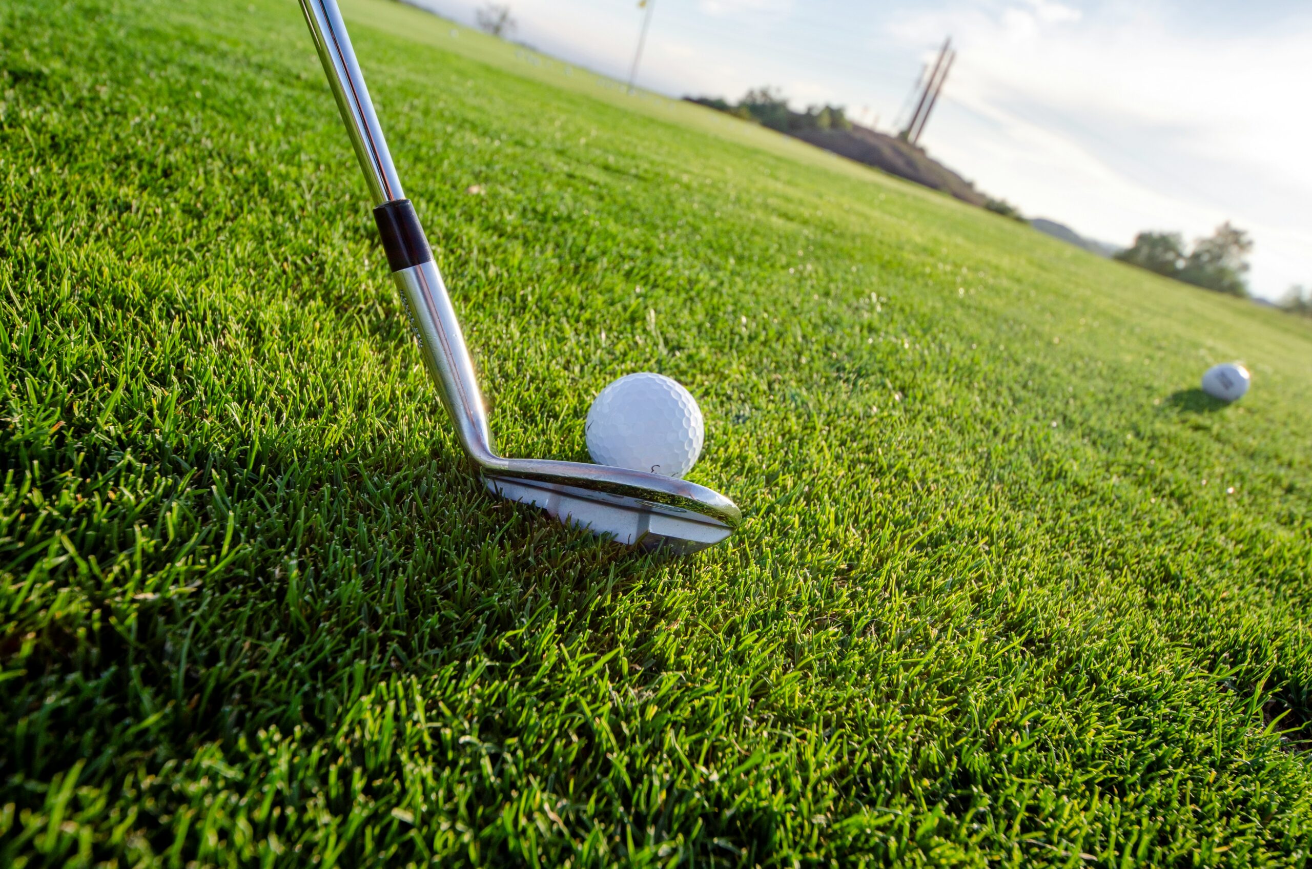 A golf club is poised to hit a white golf ball on a lush green fairway. Another ball is visible in the distance.